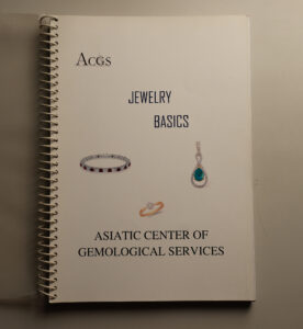 Cover of a white spiral-bound workbook titled Jewelry Basics by Asiatic Center of Gemological Services featuring images of a diamond bracelet ring and gemstone pendant